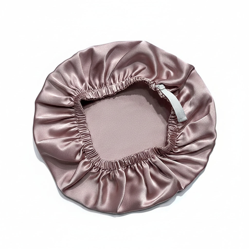 A satin sleep cap in a dusty rose pink color, laid flat on a white background. It features an elasticized opening with a white drawstring.